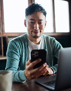 A young Asian man using his cellphone to shop car insurance from his home office, depicting how easy it is to get car insurance with GEICO. Shop auto insurance anywhere anytime.